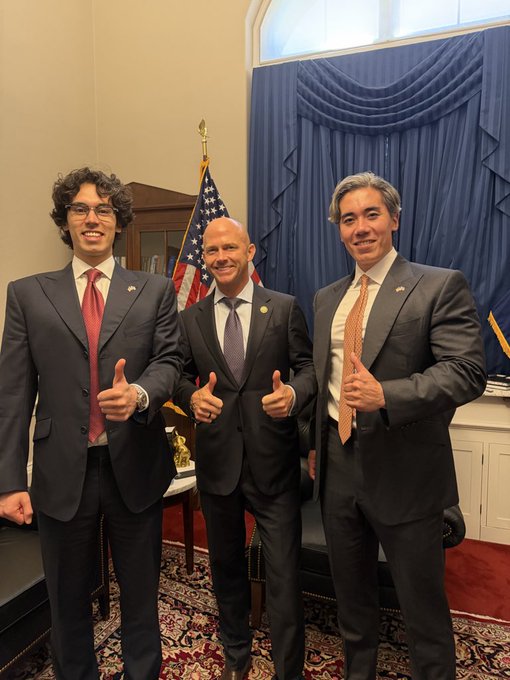 Three men in formal business suits smile and give thumbs-up gestures in what appears to be an official office with an American flag and blue draped curtains in the background.