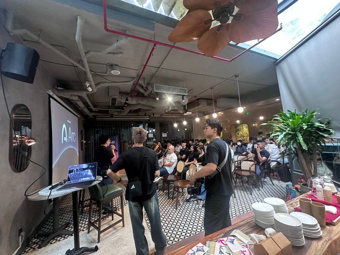 A photograph captures an indoor event space where an audience sits facing a presentation screen displaying 'Arc,' with attendees standing in the foreground and a buffet area visible on the right.