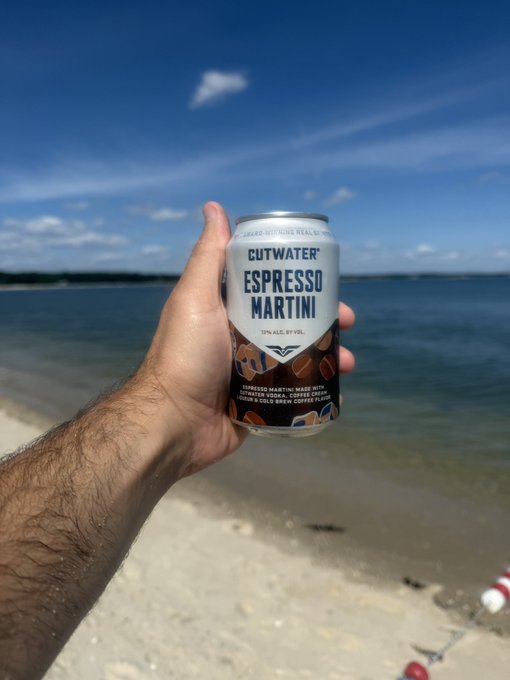 A hand holds up a can of 'Cutwater Espresso Martini' against a backdrop of a beach and ocean under a blue sky.