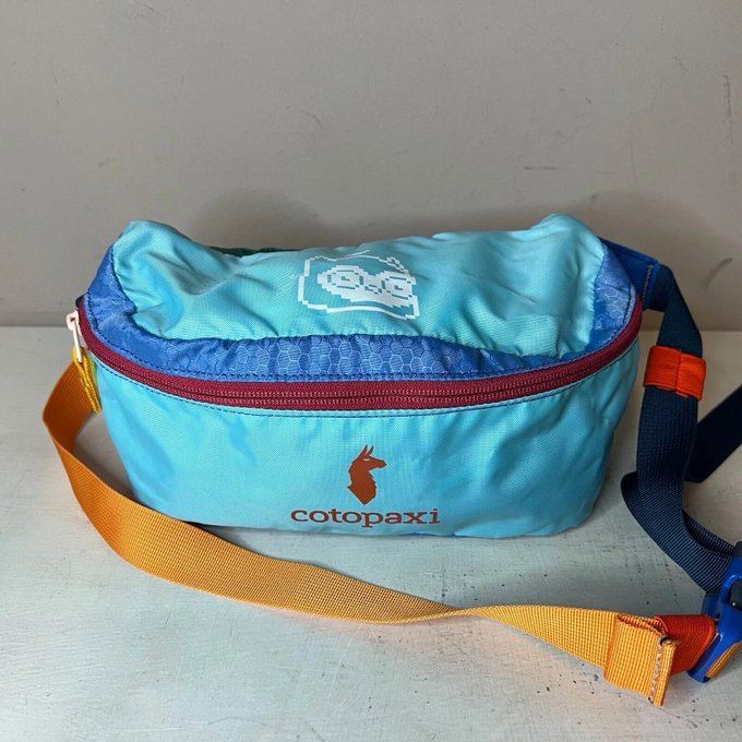 A light blue Cotopaxi fanny pack/sling bag with orange and navy straps and a pixelated logo on top, sitting against a white wall.