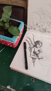 An artist's sketchbook on a green desk showing detailed pencil drawings of cats, with a black mechanical pencil resting on the open pages and a potted plant nearby.