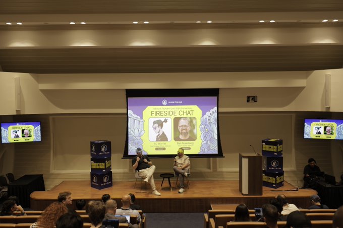 Two people sitting on a stage during a 'FIRESIDE CHAT' presentation labeled 'DRAGCON LA', with screens displaying their names and an audience visible in the foreground.