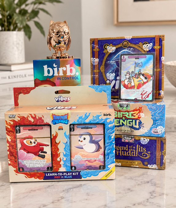 A tabletop arrangement features Pokemon Trading Card Game products including a "Learn-to-Play Kit Red vs Blue" box with Bird Trio cards visible, a "Birb in Control" promotional display with a gold owl trophy, and a "Legend of the Fuddai" booster box.