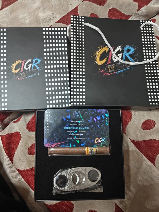 Black gift boxes branded 'CIGR' with colorful splash logos contain a CIGR Club membership card, a cigar, and a metal cigar cutter, displayed on a red and beige patterned cloth.