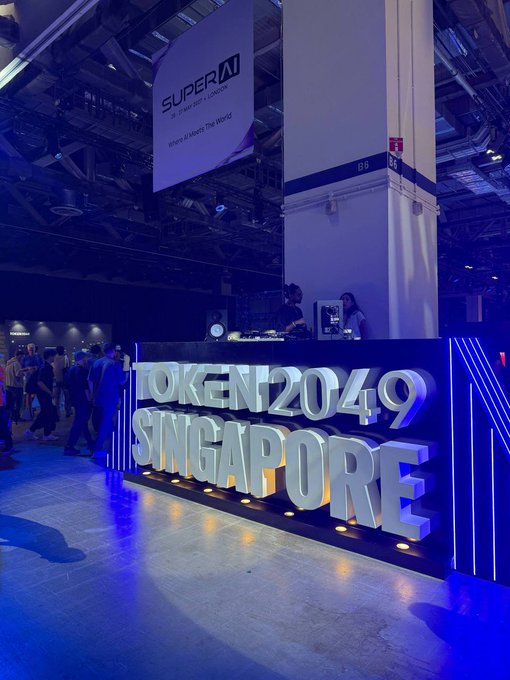An event space displays large illuminated 3D letters reading 'TOKEN2049 SINGAPORE' with a 'SUPER AI' banner hanging above in a venue bathed in blue lighting.