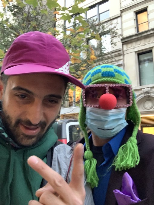 A man wearing a maroon cap and green hoodie takes a selfie with a person wearing a green knit hat with a red nose and clown-like pink glasses, who is also wearing a face mask and making a peace sign.