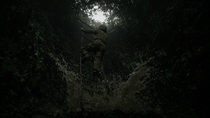 A soldier or explorer climbs a rope ladder over a muddy waterfall in a dark jungle, with splashing water rising beneath them.