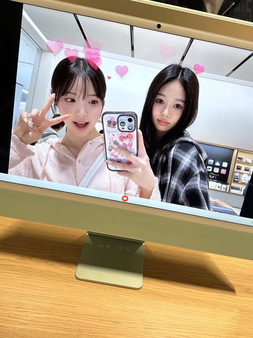 Two young women take a mirror selfie, one making a peace sign with a pink heart filter applied, while one holds a phone with a strawberry-themed case, displayed on a computer monitor.