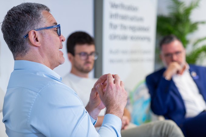 Three men sit in a discussion panel setting, with the foreground speaker wearing glasses and a light blue shirt, gesturing with his hands while two other attendees listen attentively in the blurred background.