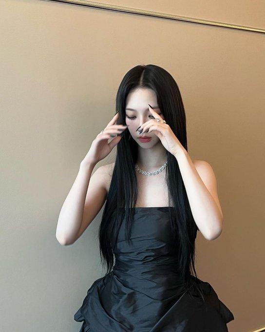 A young woman with long black hair wears a black strapless dress and a pearl necklace, posing with her hands near her face against a beige wall.