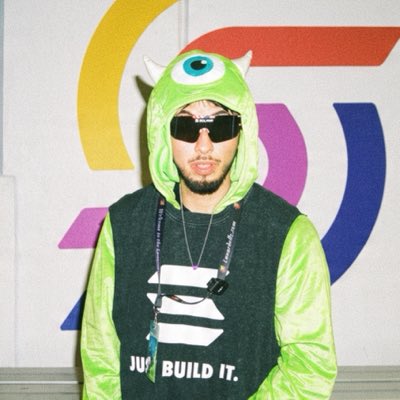 A person wears a green Mike Wazowski (Monsters Inc.) costume with sunglasses, standing in front of a colorful logo, wearing a shirt that reads 'JUST BUILD IT.'