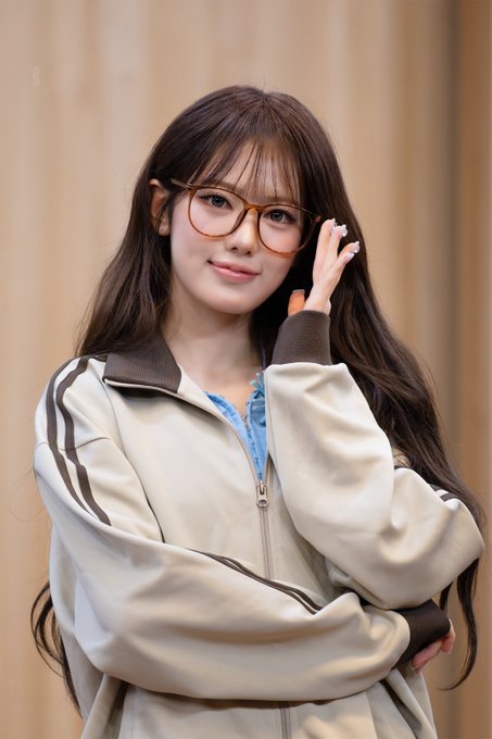 A portrait of a young woman with long brown hair, wearing oversized tortoiseshell glasses and a beige track jacket with brown stripes.