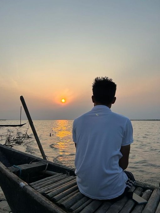 A young man in a white shirt sits on a wooden boat, watching the sun set over calm water.