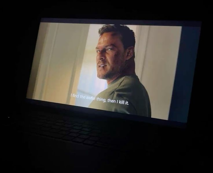 A laptop screen in a dark room playing a video showing a serious-looking man turning his head, with the subtitle 'I find the awful thing, then I kill it.'