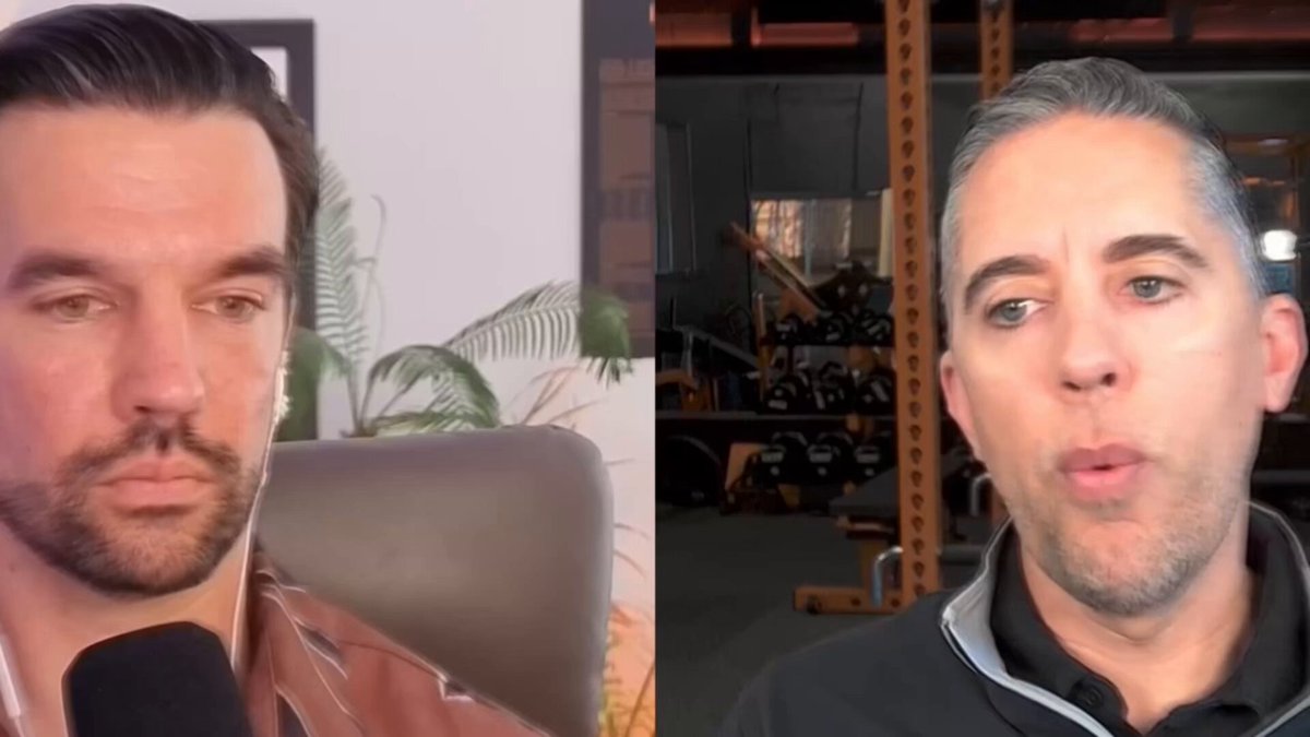 The image shows two men sitting next to each other, with one man wearing a black shirt and the other wearing a gray shirt. They are both looking at something off-camera, possibly engaged in an interview or discussion. The background of the image features a plant behind them, adding some greenery to their setting.