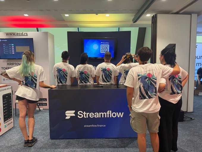 The Streamflow team, wearing matching whale-printed shirts, gathers around their branded booth at Solana Summit Serbia, engaging with attendees in front of a screen displaying the Streamflow QR code. The image captures the team's active presence at the event, aligning with their call to visit the booth and see founder @imprfekt speak on stage at 12:45 CEST.