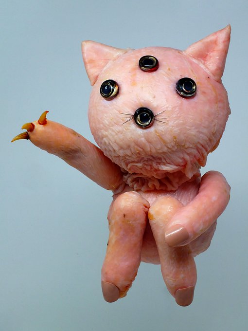 A creepy, fleshy pink cat-like figure with three eyes arranged on its face and small yellow claws sits against a plain gray background.