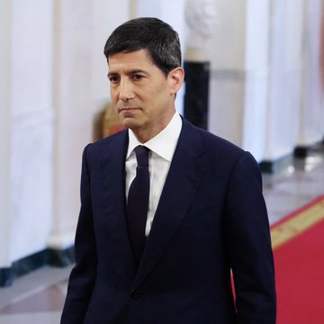 A man with short dark hair wearing a dark navy suit, white shirt, and dark tie stands in what appears to be an official government or institutional building with white columns and red carpeting visible in the background. The image is unrelated to the post about Federal Reserve interest rate predictions from Kalshi traders.