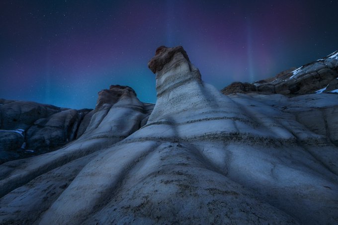 A nighttime landscape featuring tall, eroded sandstone hoodoos under a starry sky illuminated by vibrant purple and green aurora borealis.