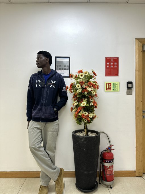 A young man wearing a navy "RXP" hoodie leans against a wall next to a tall potted plant and a red fire extinguisher. The casual indoor setting contrasts with the post's excitement about the Sui founder being in their group chat pumping the $SUI community.