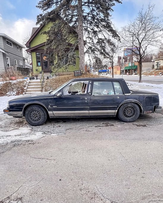 A dark sedan with broken windows is parked in front of a green house, likely a scene of vandalism that the poster is attributing to "Solana".
