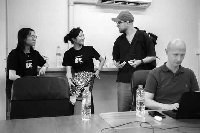 A black-and-white photograph of four people in an office or classroom setting, where two women in matching 'Wonderschool' branded t-shirts converse with a standing man wearing glasses and a cap, while another man works on a laptop at a table in the foreground.