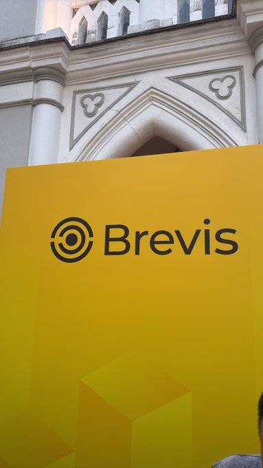 A bright yellow sign displaying the 'Brevis' logo and name in black text, mounted in front of an ornate white building with Gothic architectural details.