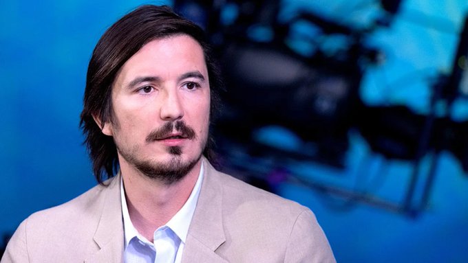 Portrait of a man with brown hair and a goatee wearing a beige suit jacket over a light blue shirt, speaking at what appears to be a conference with a blue-lit background.
