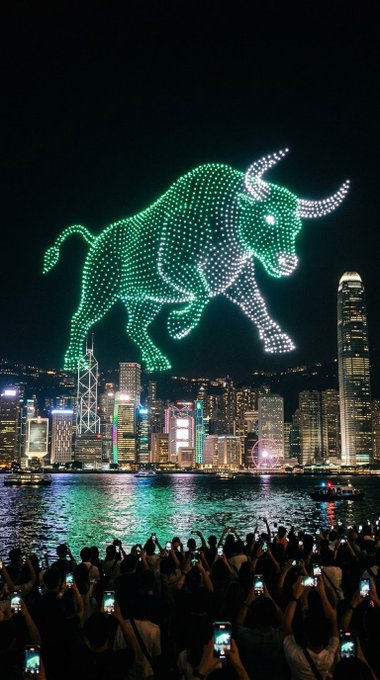 A massive green holographic bull, illuminated by hundreds of drones, charges across the night sky above Hong Kong's iconic skyline, while a crowd below captures the spectacle on their phones—a powerful "GM" (Good Morning) greeting symbolizing bullish market optimism.