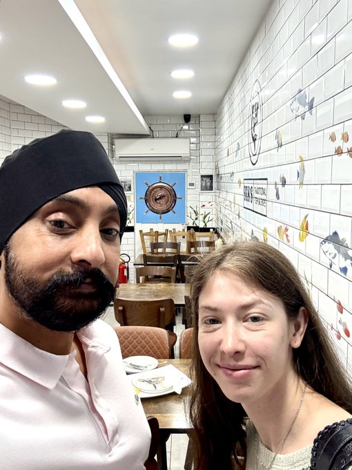 A selfie of a man wearing a black turban and a woman with long brown hair inside a restaurant featuring nautical-themed wall art.