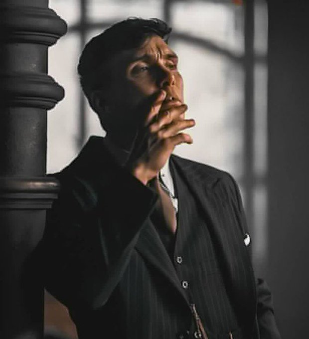 A moody cinematic portrait of a man in a dark pinstripe suit smoking a cigarette, leaning against a column with dramatic side lighting and shadows.