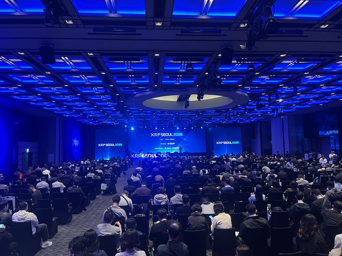 A large audience sits in a blue-lit conference hall facing a stage with screens displaying 'XRPSEOUL 2025'.