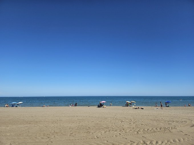 A bright sunny beach scene with clear blue skies, sandy shore, and several people with umbrellas scattered along the water's edge.