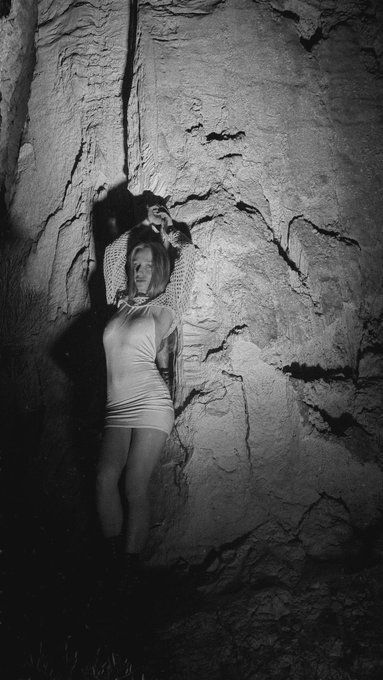 A black and white photograph shows a woman standing against a rugged rock wall in a dimly lit environment, wearing a fitted dress and boots.