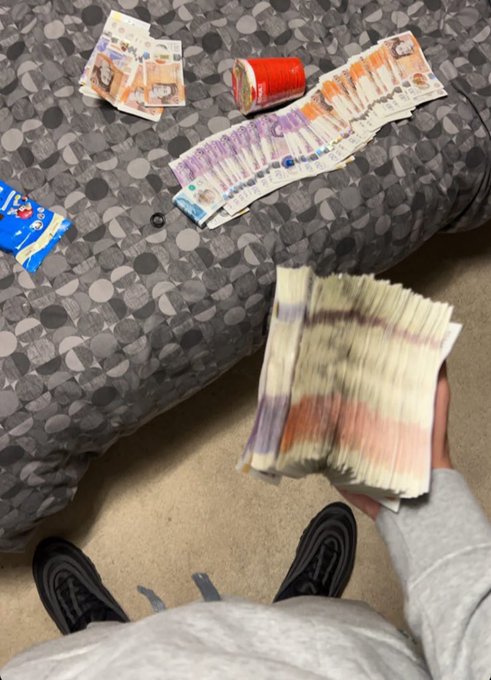 A person holds a thick fan of British pound banknotes above a bed scattered with more stacks of cash, a can, and a red container, while standing in socks. The image reflects the captioned feeling of being dazzled by an overwhelming amount of money.