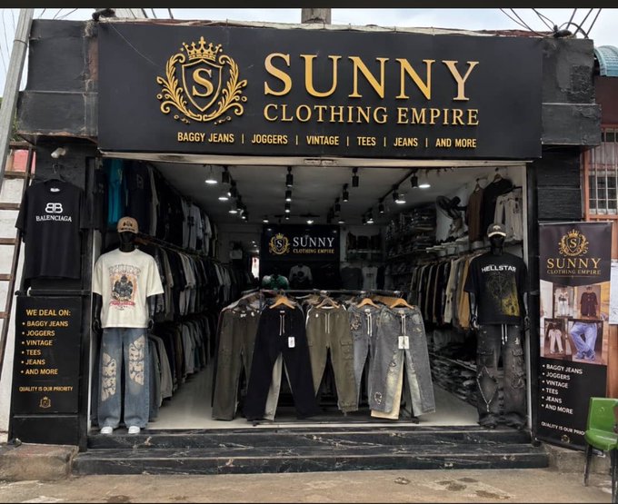 The storefront of 'SUNNY Clothing Empire' displays a black and gold sign with 'Baggy Jeans, Joggers, Vintage, Tees, Jeans, and More,' featuring mannequins dressed in casual clothing and rows of pants and shirts on racks.