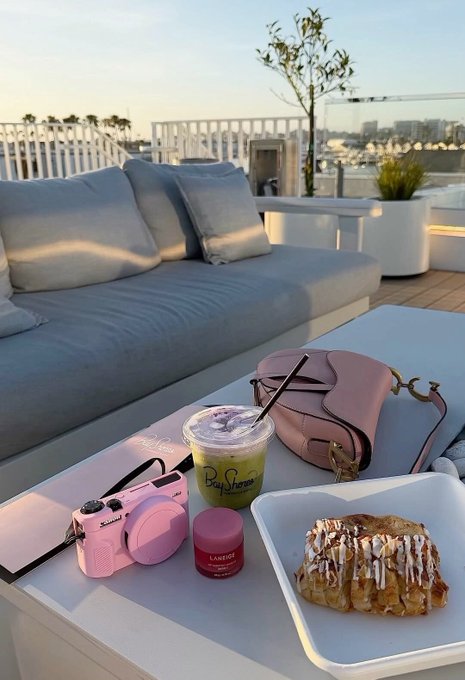 The image shows a white table with various items, including a pink camera, a cup of coffee or tea, and a pastry. The table also has a purse placed next to it. The background features an outdoor setting with a view of water and buildings, suggesting that this scene takes place in a park or similar public space. The image is related to the post because it highlights how the internet was built for classical computing eras and emphasizes the need for future innovations to be prepared for the quantum era.