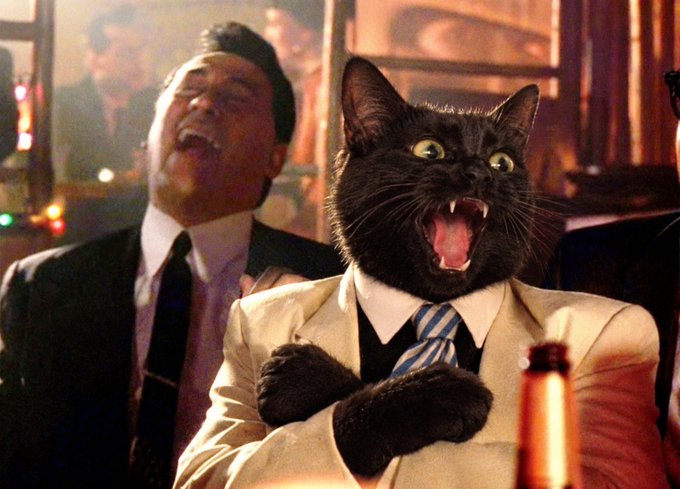 A humorous, AI-generated or edited image showing a black cat's head photoshopped onto a man in a tan suit and tie, with its mouth wide open, standing next to a laughing man in a dark suit.