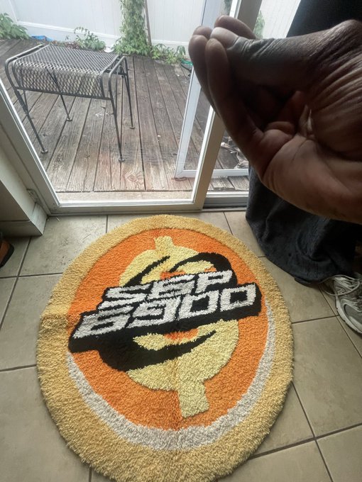 A doormat featuring a 'San Diego Padres' logo design sits on a tiled floor, with a person's hand gesturing toward it and a sliding glass door leading to a wooden deck in the background.