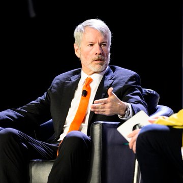 Michael Saylor, executive chairman of MicroStrategy, speaks at a conference while gesturing with his hand, likely elaborating on his unconventional Bitcoin investment strategy. The image captures him mid-discussion, visually reinforcing his commentary about favoring a BTC/USD weighting over all-in Bitcoin accumulation for long-term portfolio growth.