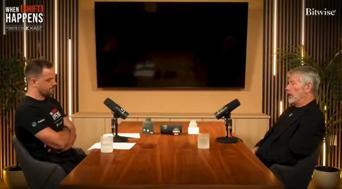 Two men converse at a wooden table, with a large flat screen TV and potted plants in the background.