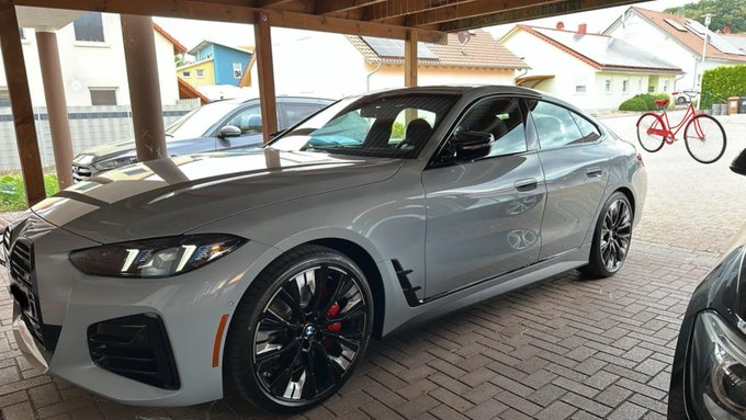 A grey BMW coupe parked under a covered structure, featuring black wheels with red brake calipers, with a red bicycle visible in the background.