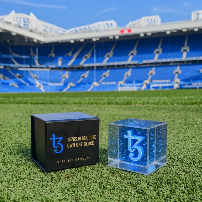 A promotional product photo for the 'Tezos Block Cube - Own One Block,' showing a black box next to a clear acrylic cube with the Tezos logo, set on a soccer field with stadium seating in the background.