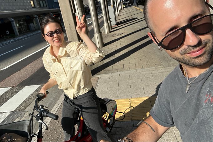 A selfie-style photo shows a man with sunglasses taking the picture while a woman in a yellow blouse waves from a bicycle on a sunny street.