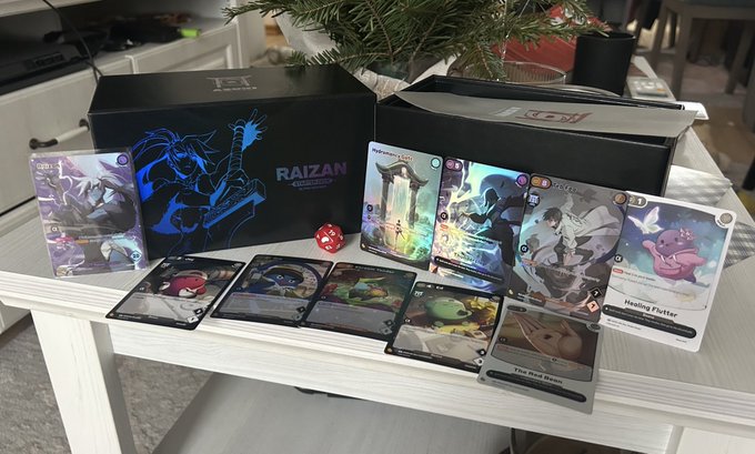 The image shows a collection of Pokémon cards displayed on a white table. The cards are neatly arranged in two rows, with some cards overlapping each other. There is also a small plant placed next to the cards, adding a touch of greenery to the scene. The text "RAIZAN" and "RAIZAN Card Game" can be seen, indicating that these cards are part of a Pokémon card game or a similar gaming experience.