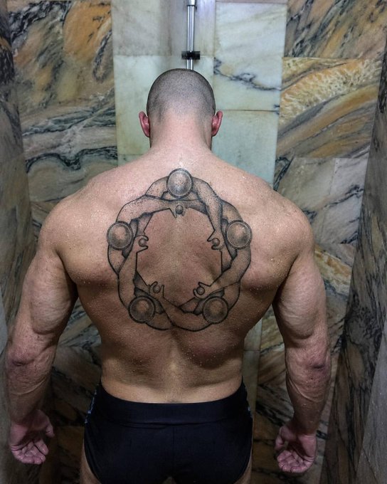 The image features a man with a large tattoo on his back, which depicts a circle of five people holding hands. The tattoo is located behind the man's shoulders and stretches across his entire back. The background of the image shows a shower stall or bathtub, suggesting that this might be taking place in a public bathroom. The caption to the image reads: "Got a tattoo in honor of @ethos_network Ethos everywhere!".
