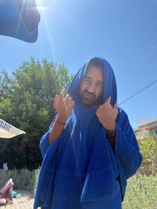 A man with a beard stands outdoors on a sunny day wrapped in a blue hooded towel or blanket, pulling the edges together and looking at the camera with an amused expression.