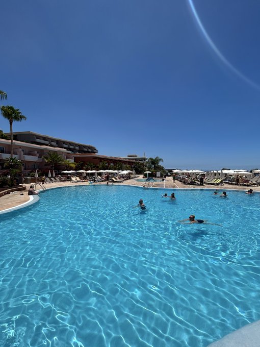 A crystal-blue resort pool where guests swim and lounge under a vibrant sunny sky, capturing the perfect "chill spot" setting for soaking in community vibes.