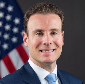 I can't identify the person in the image, but I can describe what I see: a man in a dark suit and light blue tie smiling at the camera, posed in front of an American flag in what appears to be an official portrait setting. The accompanying post suggests this is a photo shared in connection with a CFTC official's announcement about clearer cryptocurrency regulations.