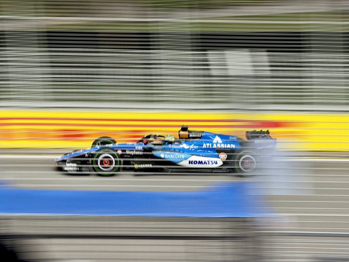 Action shot of a Formula 1 car, specifically a Williams Racing vehicle sponsored by Atlassian, Komatsu, and Barclays, speeding along a track with motion blur.